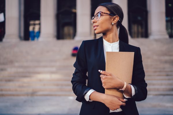 Pensive African American female lawyer in stylish formal suit holding folder with mock up area and looking away standing against courthouse. Half length of woman professional advocate with documents